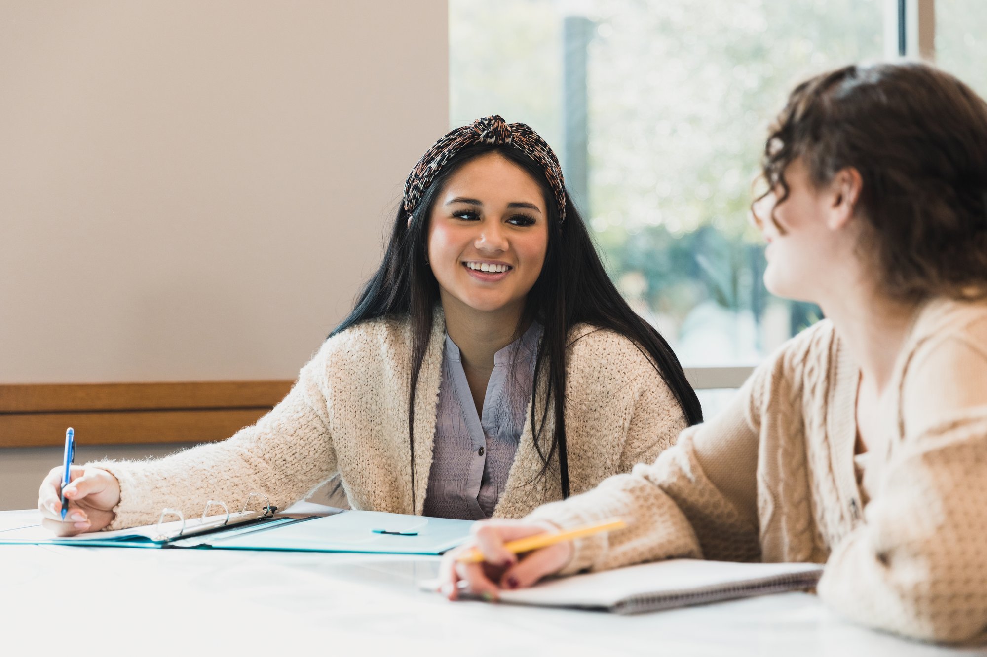 Two students at a table taking notes, looking at one another.