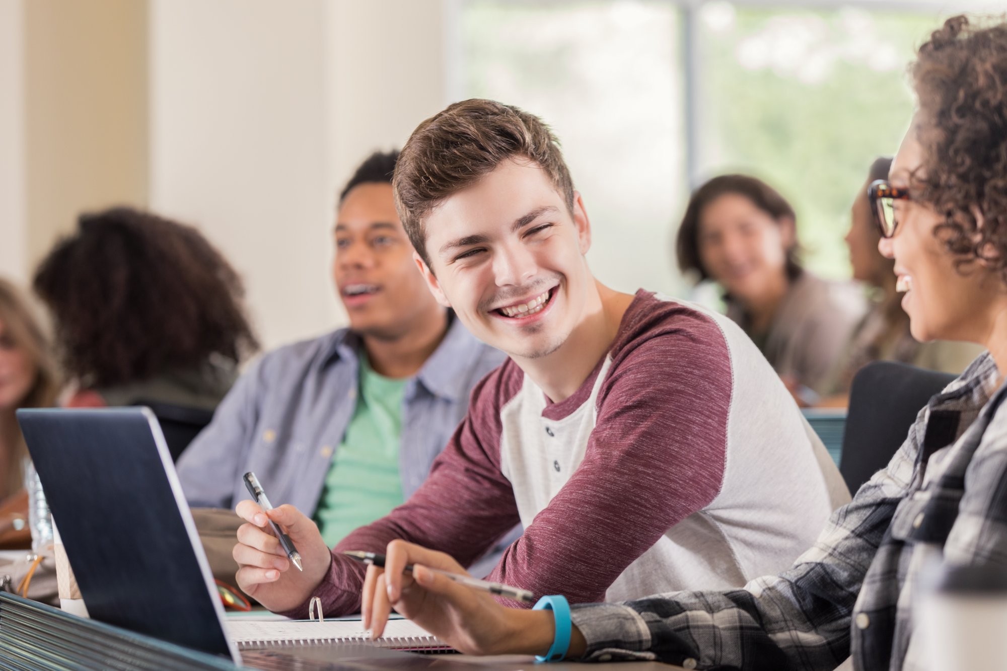 Student with pen and notebook speaking to another student on their laptop.